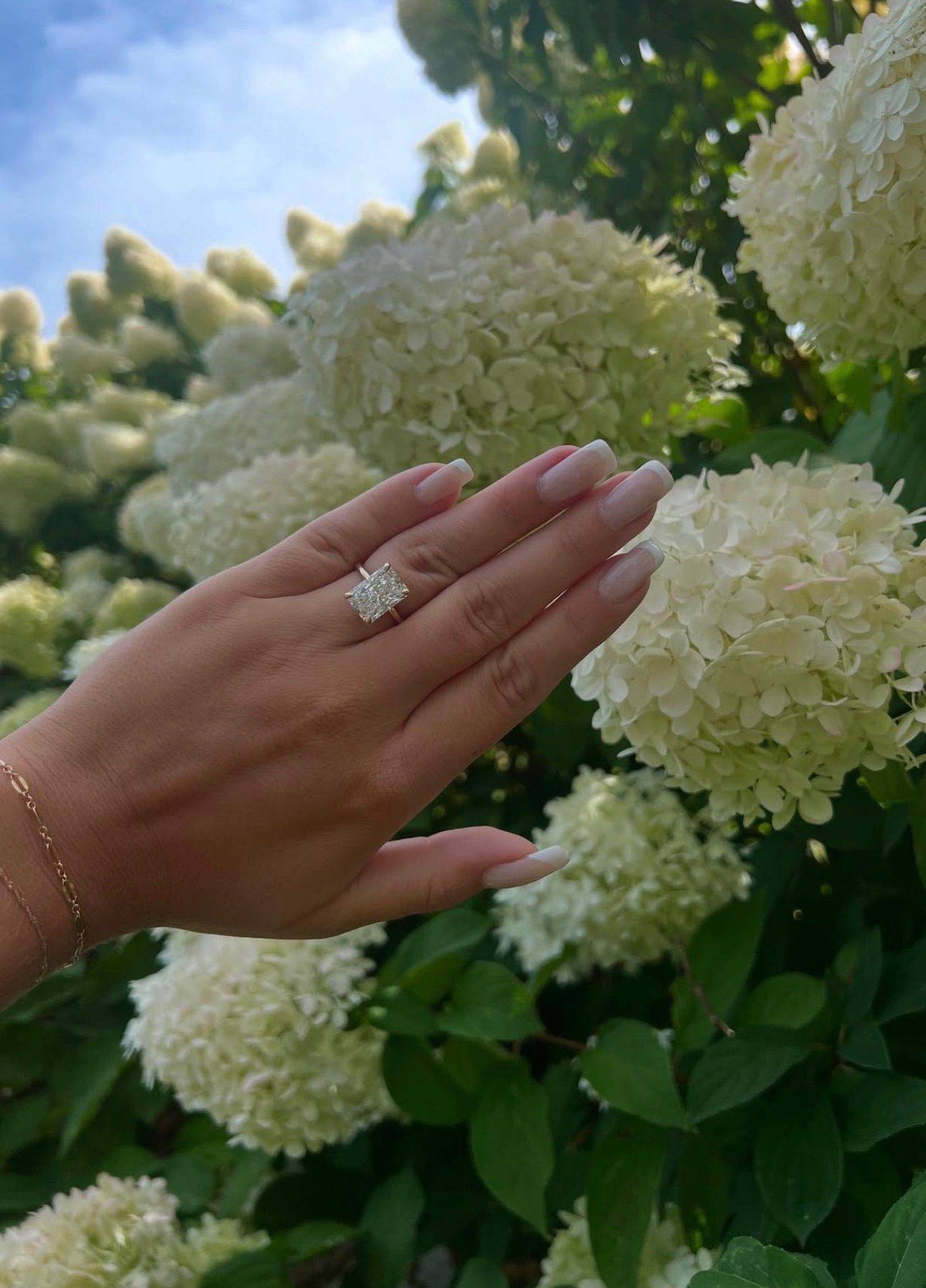 A radiant diamond ring on a pavé band from Homsy Jewelers, held against hydrangeas